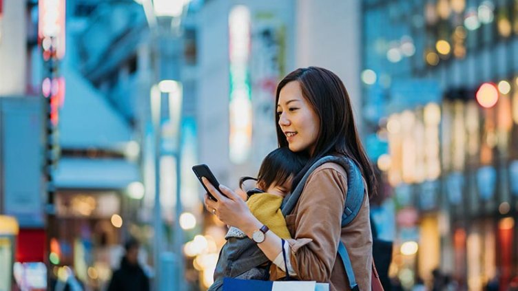 Woman with baby carrying shopping bags looks at phone while in urban retail environment 