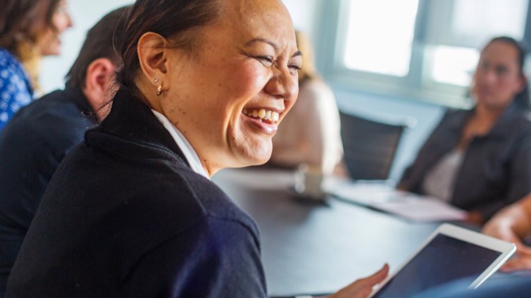A smiling woman holding a tablet sits at a conference table with a group of colleagues during a meeting.