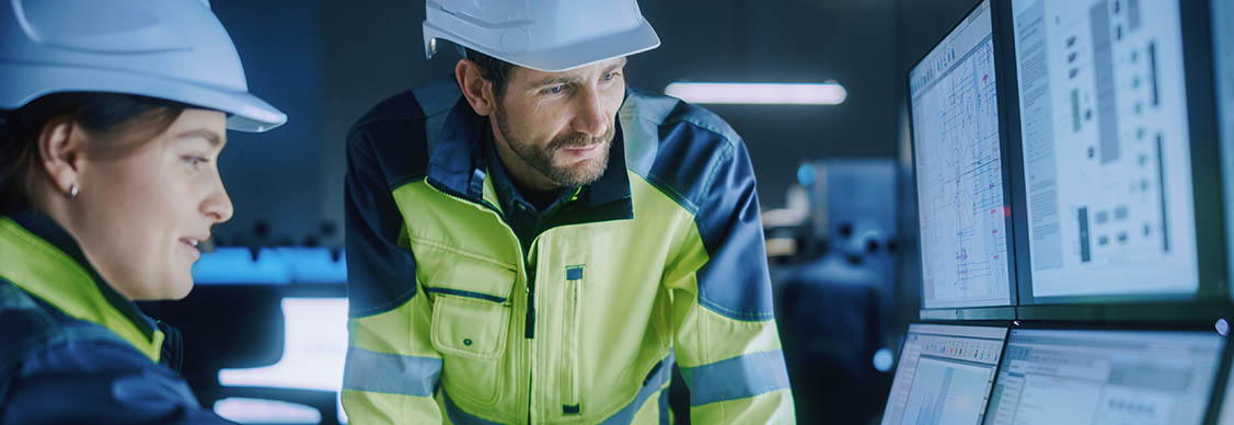 Male and female facilities managers in PPE analyzing smart building data on a bank of computer monitors