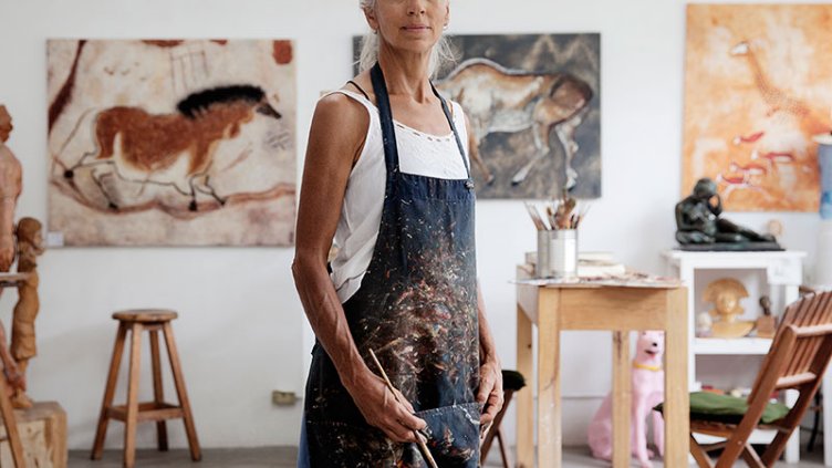 Female artist holding a paint brush standing in front of paintings